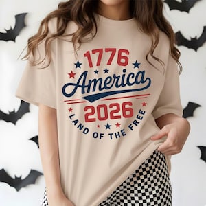 May include: Beige t-shirt with the text "1776 America 2026 Land of the Free" in red and blue. The design is circular with stars and a banner. The shirt is short-sleeved and has a relaxed fit.