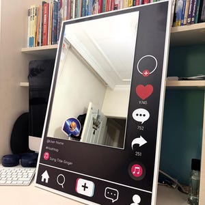 May include: A rectangular mirror with a white frame featuring a black border printed with social media interface icons, including a heart, comment bubble, share arrow, and music note, along with placeholders for a username, hashtag, and song title.