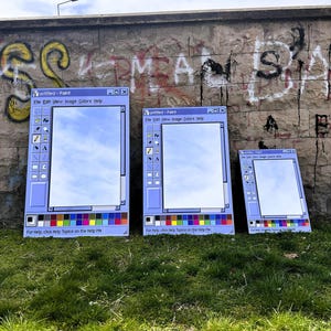 Puede incluir: Tres grandes espejos con marco azul con la forma de la interfaz del programa Paint de Windows cl&aacute;sico, que reflejan el cielo. Se exhiben al aire libre contra una pared cubierta de grafitis y hierba verde. El texto "Untitled - Paint" es visible.