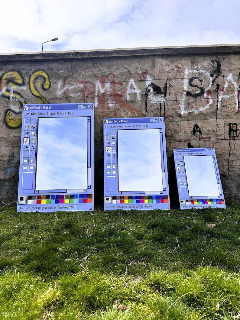 Puede incluir: Tres grandes espejos con marco azul con la forma de la interfaz del programa Paint de Windows cl&aacute;sico, que reflejan el cielo. Se exhiben al aire libre contra una pared cubierta de grafitis y hierba verde. El texto "Untitled - Paint" es visible.
