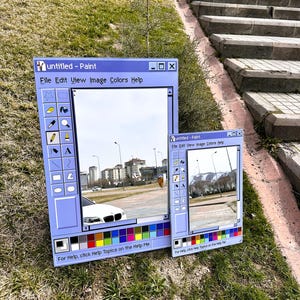 Könnte beinhalten: Zwei blaue, fensterförmige Kunstinstallationen, die an das klassische Windows Paint-Programm erinnern, sind auf Gras platziert. Die größere zeigt eine Straßenszene, während die kleinere sie spiegelt. Text enthält "untitled - Paint" und "File Edit View Image Colors Help".