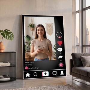 Op de afbeelding: Een groot ingelijst kunstwerk dat een social media-interface toont met een lachende persoon die een telefoon vasthoudt. De interface bevat een profiel, hashtag en songtitel. De achtergrond toont planten en een raam.