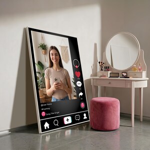 Op de afbeelding: Een groot ingelijst canvasdoek met een social media-interface met een persoon die een telefoon vasthoudt. De print leunt tegen een muur in de buurt van een kaptafel met een ronde spiegel en een roze poef.