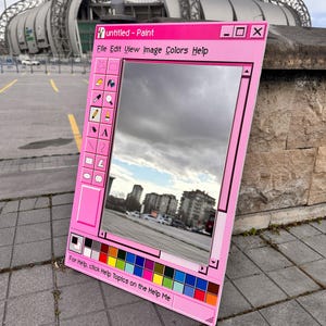 Può includere: Specchio rosa a forma di finestra Paint, che riflette un cielo nuvoloso e degli edifici. La cornice presenta la scritta "untitled - Paint" e una tavolozza di colori. Lo specchio è appoggiato su una superficie pavimentata.