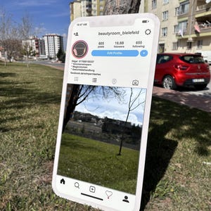 以下が含まれることがあります： スマートフォンに表示されたInstagramのプロフィール「beautyroom_bielefeld」。フォロワー数と投稿数が表示されています。スマートフォンは屋外にあり、草、木、建物の背景が見えます。表示されている時刻は午前10時47分です。