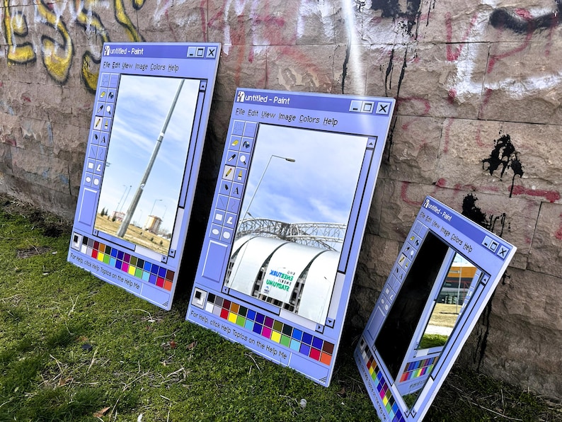 Puede incluir: Tres espejos grandes con marco azul claro apoyados contra una pared de ladrillos cubierta de graffiti. Los espejos est&aacute;n dise&ntilde;ados para parecerse a la interfaz cl&aacute;sica del programa Paint de Windows, con una paleta de colores y una barra de men&uacute;. Los reflejos muestran escenas al aire libre.