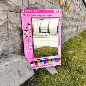 Può includere: Uno specchio rosa a forma di finestra Paint, con la scritta "untitled - Paint". Riflette un edificio ed erba. La cornice include una tavolozza di colori e icone di strumenti di disegno. Lo specchio è appoggiato su un'area erbosa.