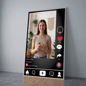 Op de afbeelding: Een ingelijste poster met een social media-interface met een vrouw die een smartphone vasthoudt. De zwarte interface bevat een profielpictogram, een hart, een commentaarbubbel en een deelpictogram. De poster heeft een witte rand en leunt tegen een grijze muur.