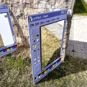 Puede incluir: Dos grandes marcos rectangulares de color azul claro que imitan la interfaz del programa Paint de Windows. Los marcos est&aacute;n apoyados sobre hierba contra un muro de piedra. El texto "untitled - Paint" es visible en la parte superior, junto con otras opciones de men&uacute; e iconos.