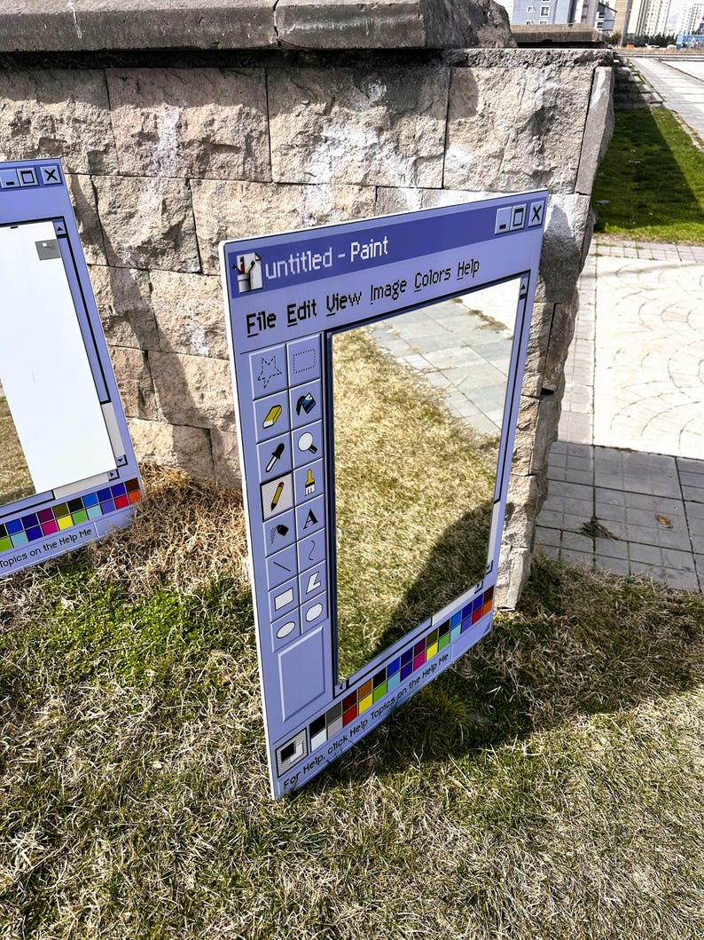 Puede incluir: Dos grandes marcos rectangulares de color azul claro que imitan la interfaz del programa Paint de Windows. Los marcos est&aacute;n apoyados sobre hierba contra un muro de piedra. El texto "untitled - Paint" es visible en la parte superior, junto con otras opciones de men&uacute; e iconos.