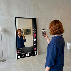 Op de afbeelding: Een grote, rechthoekige spiegel met een wit frame, ontworpen om op een social media-interface te lijken. De spiegel reflecteert een persoon die een foto maakt met een smartphone. De interface bevat pictogrammen voor likes, reacties en shares, evenals tekstvelden.