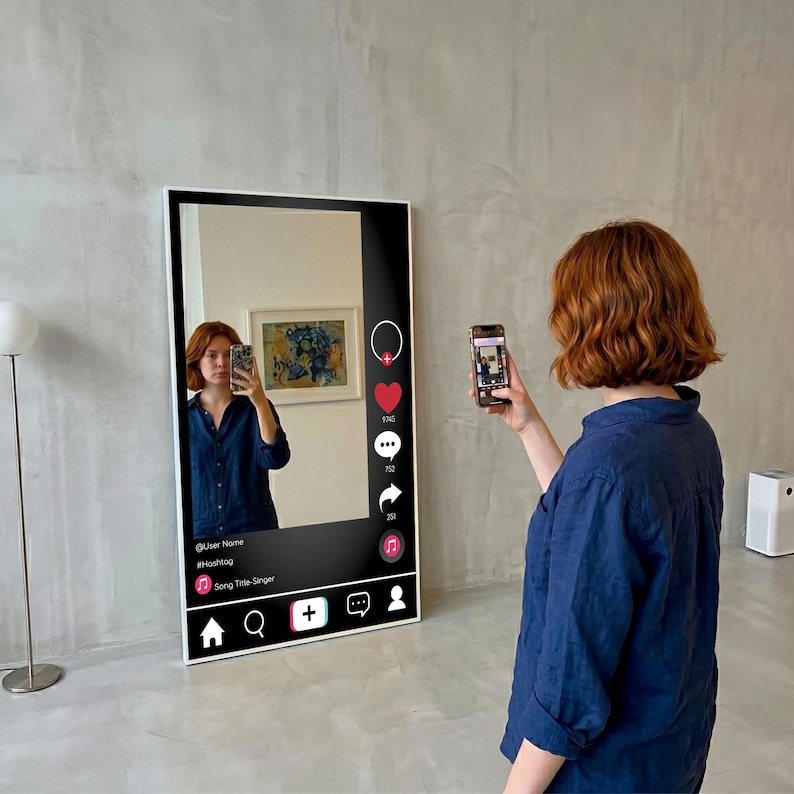 Op de afbeelding: Een grote, rechthoekige spiegel met een wit frame, ontworpen om op een social media-interface te lijken. De spiegel reflecteert een persoon die een foto maakt met een smartphone. De interface bevat pictogrammen voor likes, reacties en shares, evenals tekstvelden.