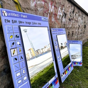 Puede incluir: Tres grandes espejos enmarcados en azul claro apoyados contra una pared de piedra, cada uno parecido a una ventana cl&aacute;sica de Paint de Windows. Los espejos reflejan un paisaje urbano y el entorno, con la barra de t&iacute;tulo "untitled - Paint" visible.