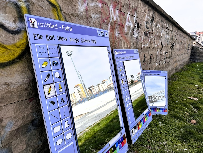 Puede incluir: Tres grandes espejos enmarcados en azul claro apoyados contra una pared de piedra, cada uno parecido a una ventana cl&aacute;sica de Paint de Windows. Los espejos reflejan un paisaje urbano y el entorno, con la barra de t&iacute;tulo "untitled - Paint" visible.