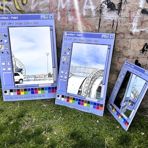 Puede incluir: Tres obras de arte rectangulares de color azul claro que imitan la interfaz de Windows Paint, cada una mostrando una escena exterior diferente. Las obras de arte est&aacute;n apoyadas sobre hierba contra una pared de ladrillo cubierta de graffiti. El texto "Untitled - Paint" es visible.