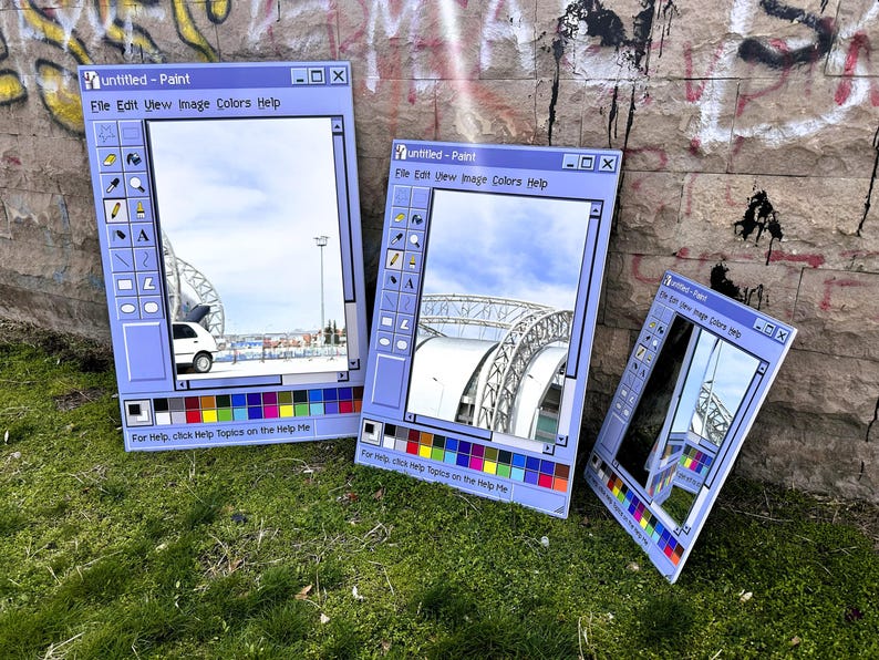 Puede incluir: Tres obras de arte rectangulares de color azul claro que imitan la interfaz de Windows Paint, cada una mostrando una escena exterior diferente. Las obras de arte est&aacute;n apoyadas sobre hierba contra una pared de ladrillo cubierta de graffiti. El texto "Untitled - Paint" es visible.