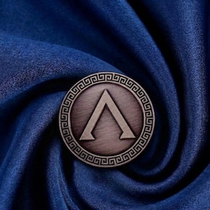 Peut inclure: Une épingle ronde en métal de couleur bronze, ornée d'une bordure grecque et d'un symbole lambda stylisé au centre. L'épingle est posée sur un fond de tissu bleu profond et fluide.