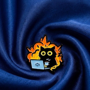Könnte beinhalten: Ein schwarzer Katzen-Emaille-Pin mit gelben Augen, der an einem Laptop mit einem Fischsymbol arbeitet und von orangefarbenen Flammen umgeben ist. Die Katze hält eine Kaffeetasse. Der Pin befindet sich auf einem dunkelblauen, strukturierten Hintergrund.
