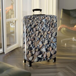 Könnte beinhalten: Ein Rollkoffer mit einem Design aus Kieselsteinen und Sand. Der Koffer hat einen schwarzen Griff und Räder. Das Design verleiht dem Koffer eine natürliche Ästhetik. Der Koffer steht auf einem Holzboden. Der Koffer ist etwa 70 cm hoch.