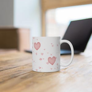 Könnte beinhalten: Weiße Keramiktasse mit rosa Aquarellherzen in verschiedenen Größen. Die Tasse hat einen gebogenen Henkel und steht auf einer Holzoberfläche. Im Hintergrund ist ein Laptop zu sehen.