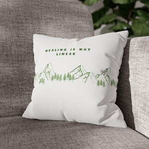 Könnte beinhalten: Weißes quadratisches Kissen mit grünem Text "HEALING IS NOT LINEAR". Darunter eine grüne Strichzeichnung von Bergen und Bäumen. Das Kissen liegt auf einem grauen Polstersessel.