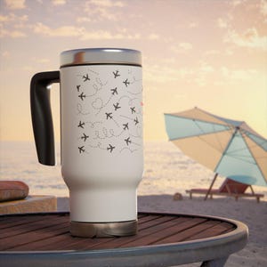 Könnte beinhalten: Ein weißer Reisebecher mit schwarzem Griff und silbernem Deckel. Der Becher hat ein Muster aus kleinen schwarzen Flugzeugen mit gepunkteten Flugrouten. Der Becher steht auf einem Holztisch in der Nähe eines Strandes mit einem Sonnenschirm.