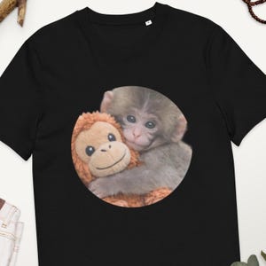 Könnte beinhalten: Schwarzes T-Shirt mit einem kreisförmigen Aufdruck eines grauen Babyaffen, der einen braunen Stofforang-Utan umarmt. Das Stofftier hat ein lächelndes Gesicht. Der Affe hat einen süßen Ausdruck. Das Shirt ist aus weichem Material.