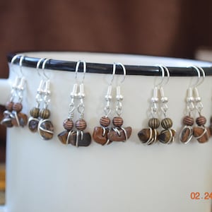 以下が含まれることがあります： 白いマグカップに飾られた、手作りのイヤリングのコレクション。イヤリングは、シルバーのフック、ワイヤーで包まれた茶色の石、木製のビーズが特徴です。「02.24.2020」の日付がマグカップに表示されています。