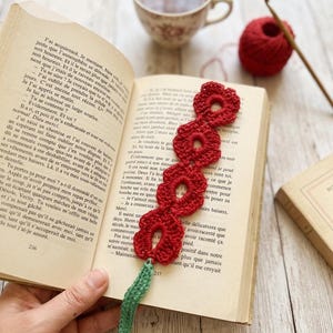 Peut inclure: Marque-page rouge en crochet avec un motif floral, posé dans un livre ouvert. Le marque-page a une tige verte. Une tasse de thé et une pelote de laine rouge sont également visibles.