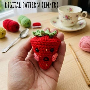 Peut inclure: Une fraise rouge au crochet avec un dessus vert et des yeux noirs est tenue dans une main. La fraise a des joues roses et une expression surprise. L'image montre également de la laine, une tasse à thé et le texte "DIGITAL PATTERN (EN/FR)".