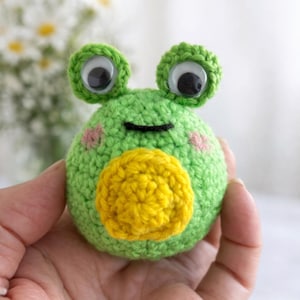 Peut inclure: Une grenouille verte au crochet avec de grands yeux globuleux, une bouche jaune et des joues roses. La grenouille est ronde et tenue dans une main, mettant en valeur la couture détaillée et le design fantaisiste. Un jouet charmant.
