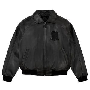 May include: A black leather bomber jacket featuring a classic collar, a full-length front zip, and ribbed knit cuffs and hem. The jacket includes two side pockets and a black embroidered emblem on the left chest with the text Avirex.
