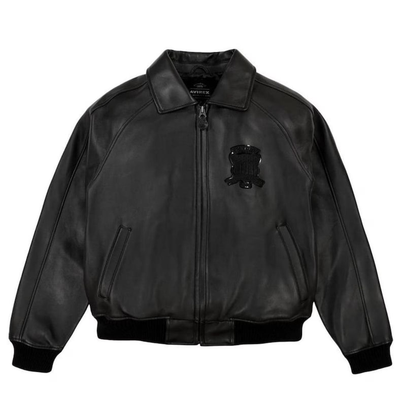 May include: A black leather bomber jacket featuring a classic collar, a full-length front zip, and ribbed knit cuffs and hem. The jacket includes two side pockets and a black embroidered emblem on the left chest with the text Avirex.