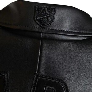 May include: A black leather jacket featuring a smooth texture and a prominent collar. Centered on the back of the collar is a black embroidered shield-shaped patch with a stylised graphic design. The jacket includes large black lettering at the bottom.