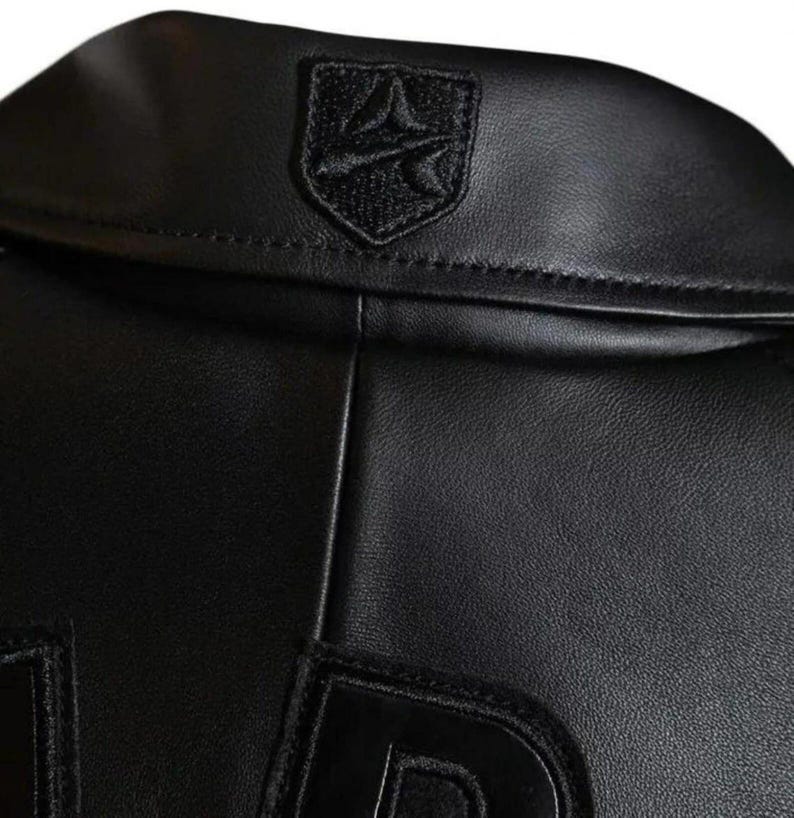 May include: A black leather jacket featuring a smooth texture and a prominent collar. Centered on the back of the collar is a black embroidered shield-shaped patch with a stylised graphic design. The jacket includes large black lettering at the bottom.