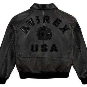 May include: A black leather bomber jacket featuring a classic collar, long sleeves with ribbed cuffs, and a ribbed hem. The back displays the embossed text AVIREX and USA in black, along with a central circular emblem featuring a graphic design.