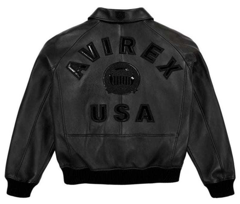 May include: A black leather bomber jacket featuring a classic collar, long sleeves with ribbed cuffs, and a ribbed hem. The back displays the embossed text AVIREX and USA in black, along with a central circular emblem featuring a graphic design.