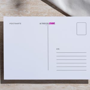 May include: A white postcard with the text "POSTKARTE" and "AUTHENTIC FAKES" in pink. A glass of amber liquid with a star anise sits to the left. A white cloth is draped on the right.