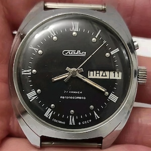 Könnte beinhalten: Eine Vintage-Armbanduhr mit schwarzem Zifferblatt, silbernem Gehäuse und römischen Ziffern. Das Zifferblatt zeigt Tag und Datum an, mit dem Markennamen "Слава". Die Uhr hat ein silbernes Armband und eine Aufzugskrone.