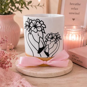 May include: A white ceramic cylindrical candle holder featuring a black line art illustration of two flamingos wearing floral crowns. The base of the holder is made of light wood and is decorated with a tied light pink satin ribbon.