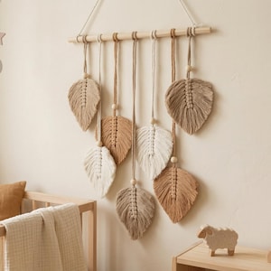 Puede incluir: Un colgante de pared de hojas de macramé en tonos beige y crema, suspendido de una varilla de madera. Las hojas están intrincadamente anudadas y cuelgan a diferentes longitudes. También se ve una cuna de madera, una almohada y un móvil con formas de luna y estrella.