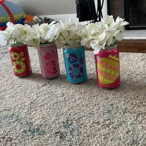 May include: Four colorful, repurposed soda cans used as vases, each holding white flowers. The cans are decorated with various designs and rhinestones around the rim. The vases are arranged on a patterned rug.