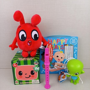 May include: A collection of children's toys, including a red plush character, a green alien figurine, a pink recorder, a coloring set, and a watermelon-themed puzzle box. The coloring set features the text "HAPPY DAYS".