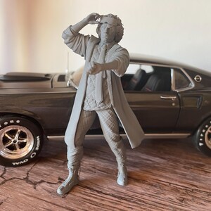 Peut inclure: Figurine grise d'un homme en long manteau et pantalon à carreaux, la main levée vers son front. Il se tient devant une voiture classique gris foncé avec des pneus noirs et des jantes chromées. La voiture est garée sur une surface en bois.
