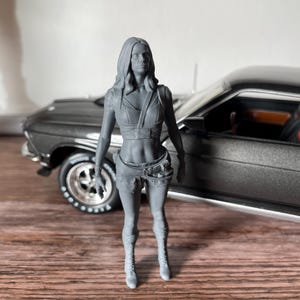 Peut inclure: Figurine grise d'une femme en crop top et short, debout devant une voiture classique grise. La voiture a des pneus noirs et un intérieur noir. La figurine porte des bottes. La voiture est sur une surface en bois.