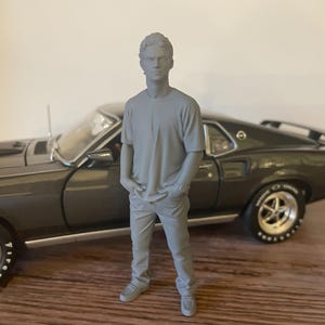 Può includere: Un modello in scala di grigi di un uomo in maglietta e pantaloni, in piedi accanto a un'auto classica grigio scuro. L'auto ha cerchi argentati e scritte bianche sui pneumatici. Il modello è in piedi con le mani in tasca.