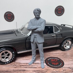 Peut inclure: Une figurine grise imprimée en 3D représentant une personne en costume, les bras croisés, debout devant une voiture miniature de type muscle car gris foncé. La voiture possède des jantes chromées, des pneus noirs Wide Oval Firestone.
