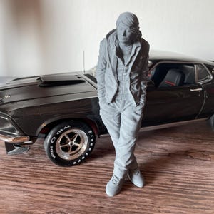 Peut inclure: Une figurine grise d'une personne en tenue décontractée se tient à côté d'une voiture classique noire. La voiture a des accents chromés et des pneus à lettrage blanc. La figurine porte une veste, un pantalon et des baskets.