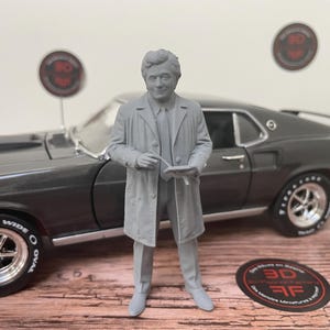 Op de afbeelding: Een grijs 3D-geprint miniatuurfiguur van een man in een trenchcoat, overhemd en das, die een klein notitieblok en een pen vasthoudt. Het figuur staat voor een donkergrijs schaalmodel van een vintage muscle car met chromen velgen en banden.