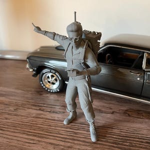 Peut inclure: Une figurine grise imprimée en 3D représentant un personnage portant une combinaison, des lunettes et un appareil de type sac à dos. Le personnage se tient debout, un bras tendu vers le haut, tenant un petit objet rectangulaire.